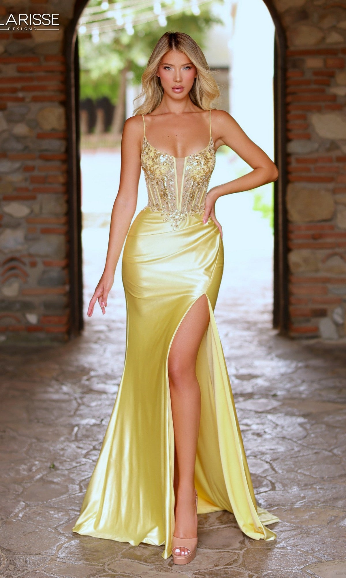A woman in a Clarisse Designs 62203 long yellow prom dress with a beaded corset bodice and high slit stands in an arched stone doorway, looking at the camera.